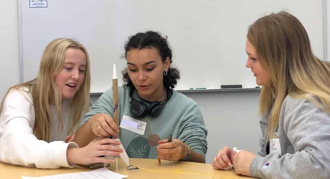 Three high school students sitting at a table assemble a small cardboard rocket 