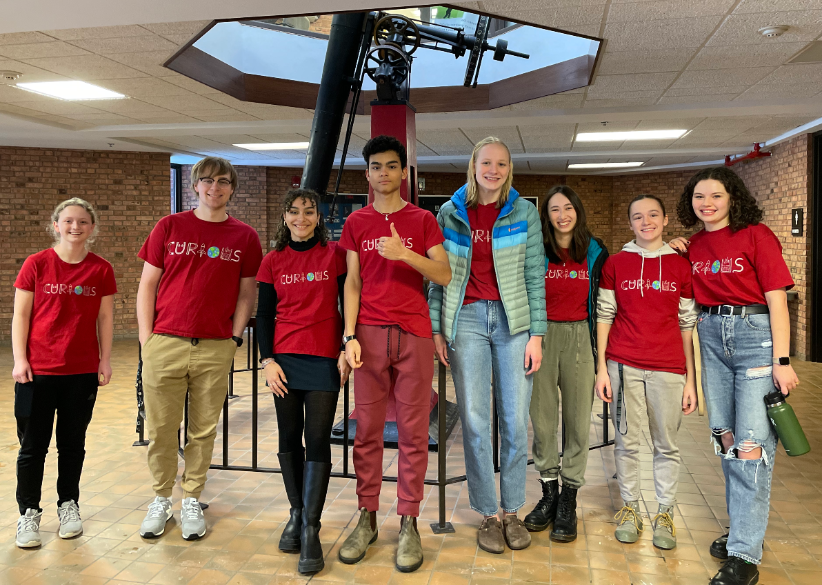 Eight of the 9 high school Teen Leaders in matching red shirts that read "Curious" standing together in front of a large telescope