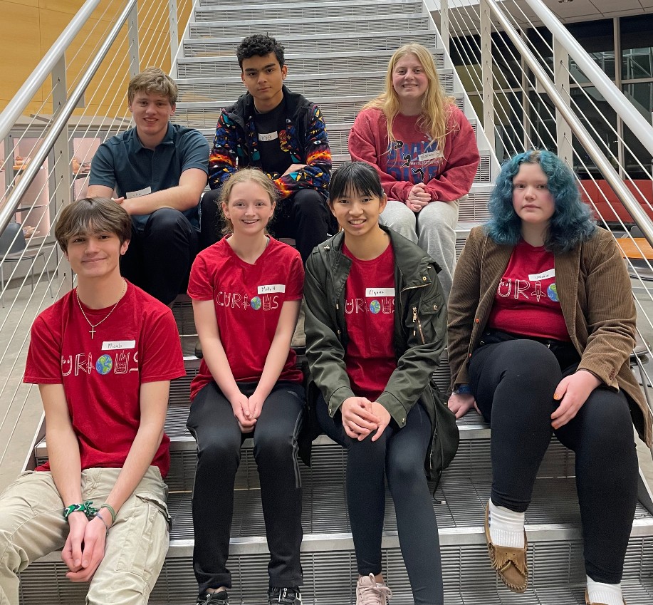 High school students serving as Teen Leaders sit on the steps in the Swenson Science Building. Four Teens sit on the lower step and three teens sit on the upper step. Four Teen Leaders are wearing matching red shirts with the word "Curious" written with STEM graphics. 